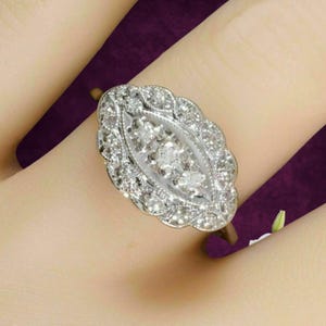 May include: A marquise-shaped ring crafted from silver-toned metal, featuring a central row of three small round-cut clear stones surrounded by a scalloped border of numerous tiny pavé-set clear stones, creating a detailed cluster design.