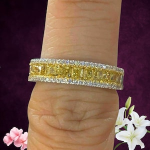 Könnte beinhalten: Ein goldener Diamantring mit einem Band aus gelben Diamanten, das von einer Reihe kleinerer, klarer Diamanten umrandet ist. Der Ring wird auf einem Finger vor einem dunkelvioletten Hintergrund mit Blumen im Vordergrund gezeigt.