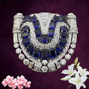 Könnte beinhalten: Silberfarbene Brosche mit symmetrischem Design, mit einer zentralen Anordnung aus ovalen und rechteckigen tiefblauen Edelsteinen. Die Brosche ist außerdem mit zahlreichen klaren, rund geschliffenen Edelsteinen verziert, die einen funkelnden Effekt erzeugen. Der Hintergrund ist tiefviolett.