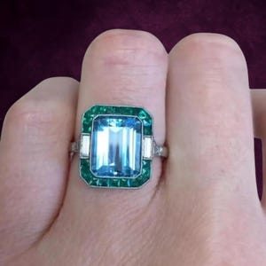 Könnte beinhalten: Rechteckiger Aquamarinring mit grünem Smaragd-Rand und weißen Akzenten. Der Ring ist auf einem silbernen Band gefasst. Der große, hellblaue Mittelstein ist der Blickfang des Rings.