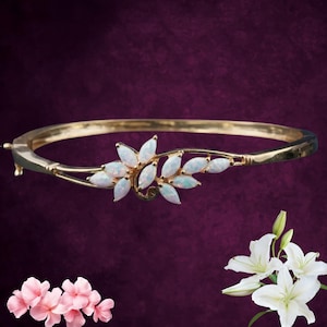 Peut inclure: Bracelet jonc doré orné d'un motif floral avec des pierres blanches semblables à des opales. Le bracelet est doté d'un fermoir à charnière et d'une esthétique délicate inspirée de la nature. Les pierres sont ovales et disposées en forme de feuilles.