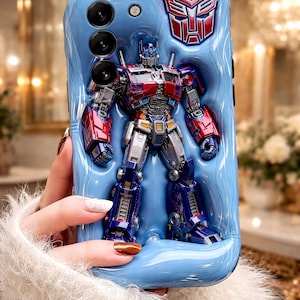 Op de afbeelding: Blauwe telefoonhoes met een 3D-ontwerp van Optimus Prime. De hoes heeft een verhoogd reliëf van de robot met rode, blauwe en zilveren details. Het Autobot-symbool is zichtbaar in de rechterbovenhoek.