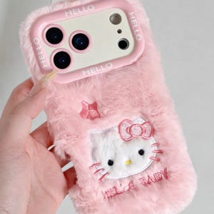Puede incluir: Funda de teléfono rosa y esponjosa con un diseño de Hello Kitty. La funda tiene un marco rosa con la palabra "HELLO" impresa. El diseño de Hello Kitty está bordado con un lazo y las palabras "HELLO BABY".