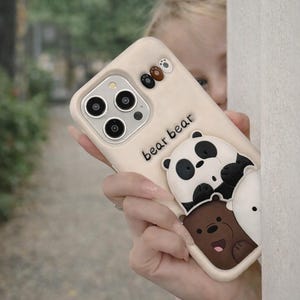 May include: A beige phone case with the words "bear bear" and a 3D design of three cartoon bears. The bears are panda, brown, and white. The phone case has a camera cutout and small bear paw prints.