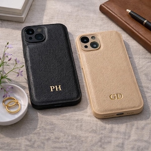 Puede incluir: Dos fundas de teléfono personalizadas. Una es negra con las iniciales doradas "PH", la otra es beige con las iniciales doradas "GD". Ambas fundas tienen un aspecto de cuero texturizado. Las fundas están sobre una superficie neutra con un plato pequeño de anillos dorados.