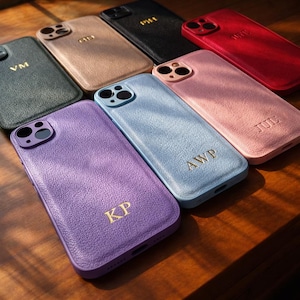 Può includere: Una collezione di cover per telefono testurizzate in vari colori, tra cui viola, azzurro, rosa, rosso, nero, beige e verde scuro. Ogni cover presenta un ritaglio per la fotocamera e iniziali dorate come KP, AWP, JUL, VM e CD.