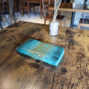 Op de afbeelding: Een turquoise en goudkleurige portemonnee met slangenpatroon ligt op een houten tafel. De portemonnee heeft een ritssluiting en een getextureerd oppervlak. De achtergrond omvat een vaas met gedroogde planten en een vage restaurantomgeving.