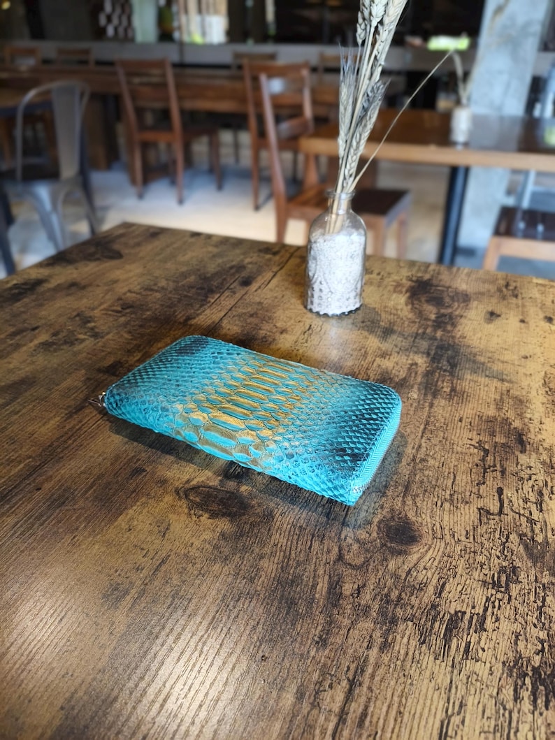 Op de afbeelding: Een turquoise en goudkleurige portemonnee met slangenpatroon ligt op een houten tafel. De portemonnee heeft een ritssluiting en een getextureerd oppervlak. De achtergrond omvat een vaas met gedroogde planten en een vage restaurantomgeving.