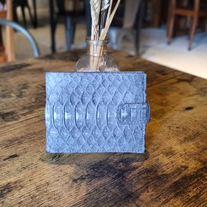 May include: A gray, textured wallet with a reptile-like pattern. The wallet is rectangular with a clasp closure. A small glass vase with dried wheat stalks sits behind the wallet on a wooden surface.