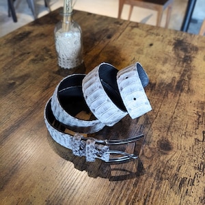 May include: A coiled, light grey belt with a reptile skin pattern. The belt has a silver-coloured buckle and a black interior. The belt is displayed on a wooden table.