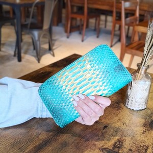 Op de afbeelding: Een turquoise en goudkleurige portemonnee met slangenpatroon en ritssluiting. De portemonnee wordt op een houten tafel gehouden. De kleur van de portemonnee is een levendig turquoise met gouden accenten.