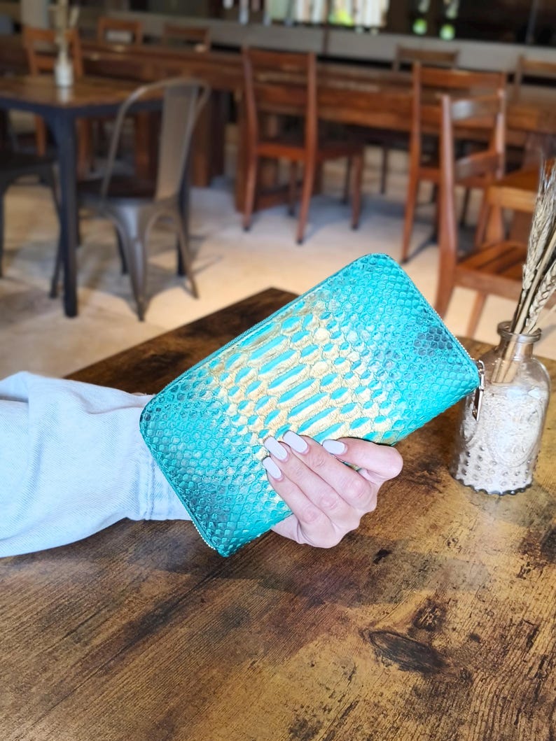 Op de afbeelding: Een turquoise en goudkleurige portemonnee met slangenpatroon en ritssluiting. De portemonnee wordt op een houten tafel gehouden. De kleur van de portemonnee is een levendig turquoise met gouden accenten.