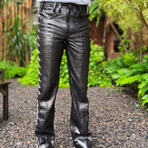 Peut inclure: Pantalon noir en similicuir imitation peau de serpent avec des accents blancs sur les côtés. Le pantalon présente un motif texturé et une conception classique à cinq poches. Le pantalon est associé à des chaussures à enfiler grises.