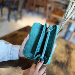 Op de afbeelding: Open turquoise portemonnee met meerdere compartimenten en een ritssluiting. De portemonnee is opengehouden en onthult het interieur. De achtergrond is wazig, met een houten tafel en een vleugje caf&eacute;sfeer.