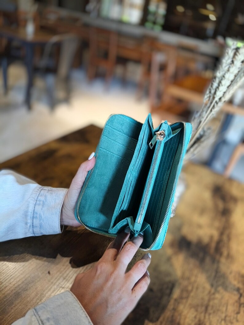 Op de afbeelding: Open turquoise portemonnee met meerdere compartimenten en een ritssluiting. De portemonnee is opengehouden en onthult het interieur. De achtergrond is wazig, met een houten tafel en een vleugje caf&eacute;sfeer.