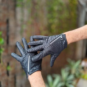 Op de afbeelding: Een paar zwarte leren handschoenen met een slangenleerpatroon. De handschoenen hebben ritsen op de polsen en worden getoond tegen een vage achtergrond van groen.