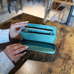 Op de afbeelding: Een open turquoise portemonnee met een gouden rits, met meerdere compartimenten. De portemonnee is gemaakt van een textuurrijk materiaal en wordt op een houten tafel getoond. De portemonnee wordt opengehouden.