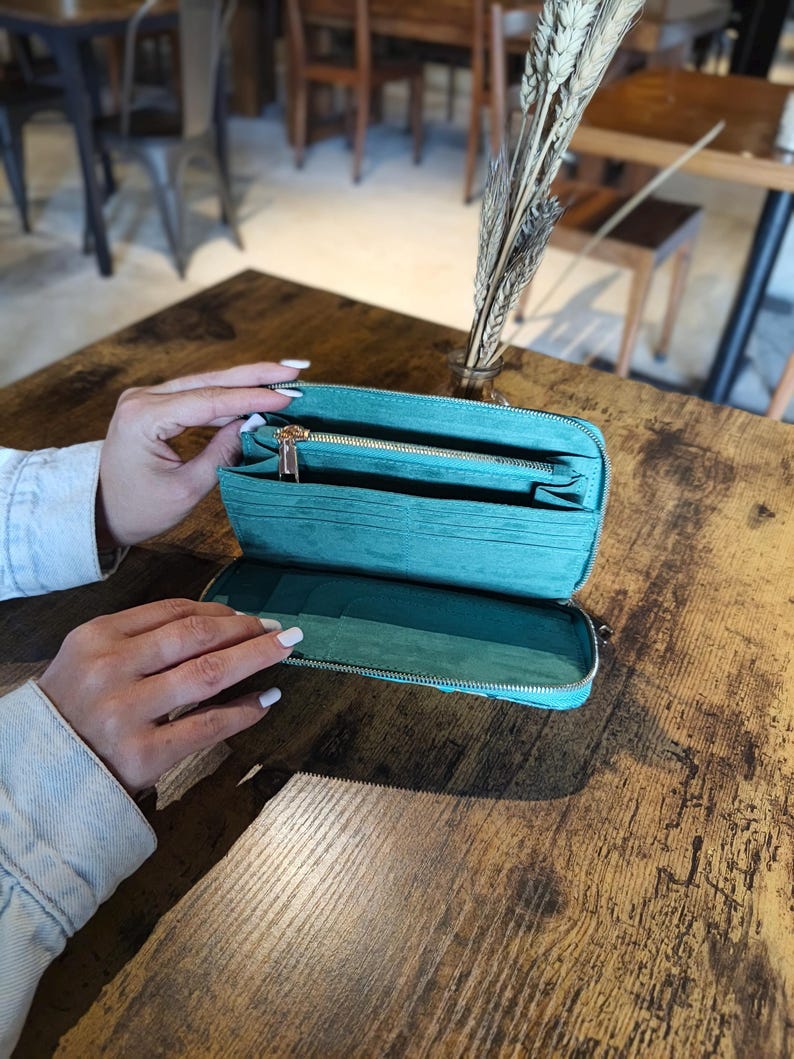 Op de afbeelding: Een open turquoise portemonnee met een gouden rits, met meerdere compartimenten. De portemonnee is gemaakt van een textuurrijk materiaal en wordt op een houten tafel getoond. De portemonnee wordt opengehouden.