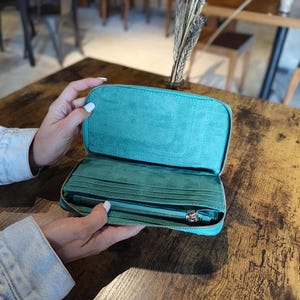 Op de afbeelding: Open teal portemonnee met meerdere kaartsleuven en een vak met ritssluiting. De portemonnee is gemaakt van een zacht, su&egrave;de-achtig materiaal en heeft een zilveren ritssluiting. De portemonnee wordt opengehouden en toont de binnenkant.