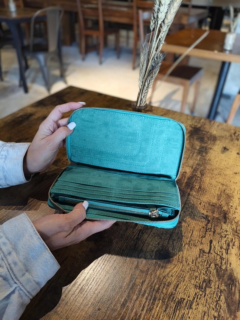 Op de afbeelding: Open teal portemonnee met meerdere kaartsleuven en een vak met ritssluiting. De portemonnee is gemaakt van een zacht, su&egrave;de-achtig materiaal en heeft een zilveren ritssluiting. De portemonnee wordt opengehouden en toont de binnenkant.