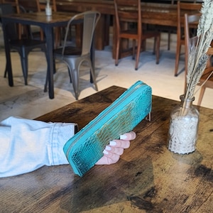Op de afbeelding: Een turquoise, rechthoekige portemonnee met een textuur van slangenleer. De portemonnee heeft een gouden rits en wordt op een houten tafel gehouden. De achtergrond toont een restaurantomgeving.