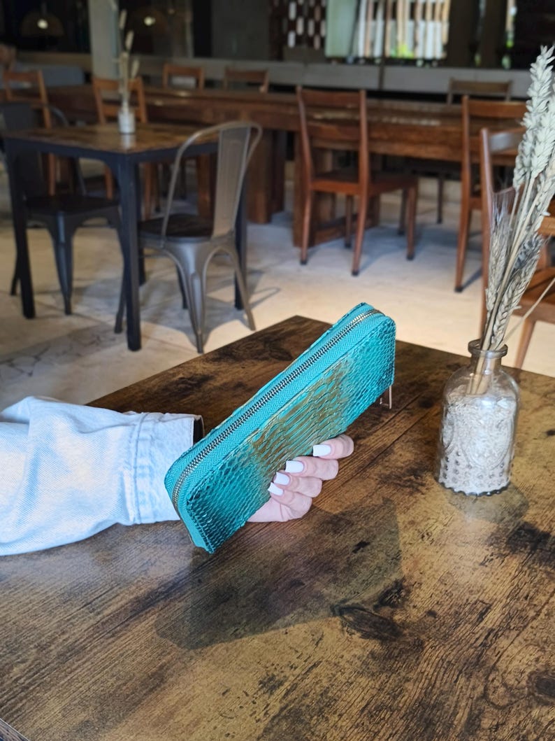 Op de afbeelding: Een turquoise, rechthoekige portemonnee met een textuur van slangenleer. De portemonnee heeft een gouden rits en wordt op een houten tafel gehouden. De achtergrond toont een restaurantomgeving.