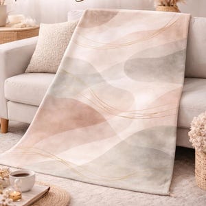 Könnte beinhalten: Eine abstrakte, neutrale Decke mit weichem, samtigem Griff. Die Decke hat ein minimalistisches Boho-Design mit fließenden Formen in Beige, Rosa und Grau, akzentuiert mit goldenen Linien. Der Text auf der Decke lautet "Abstract Neutral Blanket."