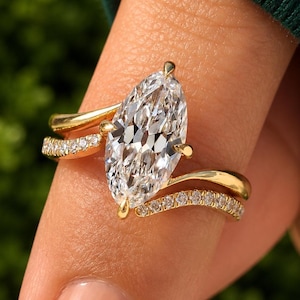 Peut inclure: Une bague en or avec une grande pierre centrale transparente taille marquise maintenue par des griffes, flanquée de deux anneaux incurvés incrustés de petites pierres transparentes rondes qui se rejoignent au centre.