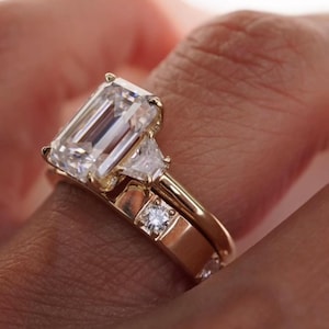 May include: Close-up of a gold engagement ring with a large emerald-cut diamond and two smaller side stones. The ring is on a finger. The text on the image reads: Have a low set engagement ring and nothing stacks flush with it?