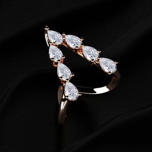 May include: A rose gold ring featuring a series of pear-shaped, clear gemstones. The gemstones are arranged in a curved, open design. The ring is set against a black background, highlighting its elegant form.