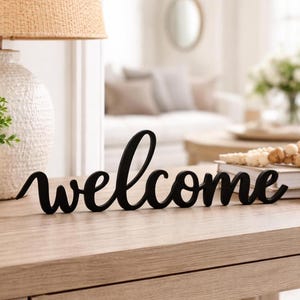 Puede incluir: Un letrero de madera negro con la palabra "welcome" escrita en cursiva. Descansa sobre una mesa de madera clara, con una lámpara, una planta en maceta y otros objetos decorativos. La escena evoca un ambiente acogedor.