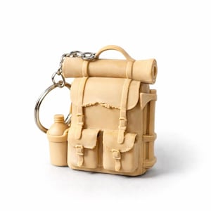 May include: A miniature beige backpack keyring with a rolled-up sleeping mat attached. The backpack has two front pockets and a small water bottle. The keyring is made of a light-coloured material and has a silver chain and ring.