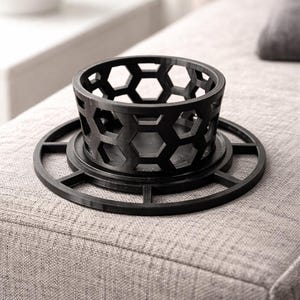 Puede incluir: Un portavelas negro con un diseño geométrico de panal. El soporte se asienta sobre una base circular con un patrón a juego. El objeto está hecho de un material oscuro y se coloca sobre una superficie tapizada gris.