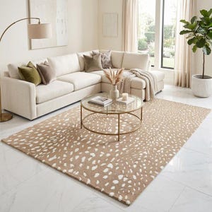 May include: A rectangular area rug featuring a tan background with a repeating white spotted pattern. The rug is positioned on a white marble floor in front of a cream-colored sectional sofa, a round glass coffee table, and a tall floor lamp.