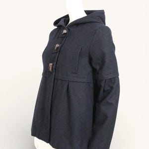 Peut inclure: Un manteau à capuche bleu marine avec un devant boutonné et un ourlet évasé. Le manteau comporte une capuche, deux poches et des fermetures à bascule en bois. Les manches ont un design unique. Le manteau est présenté sur un mannequin.