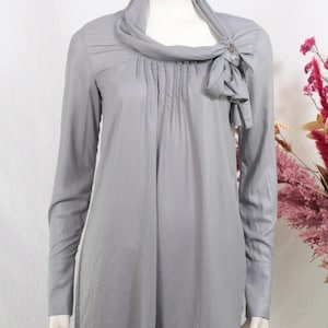 以下が含まれることがあります： ドレープの入ったハイネックと、側面に装飾的なリボンが付いた、ライトグレーの長袖ブラウス。 ブラウスは前面にプリーツのディテールがあり、マネキンに展示されています。