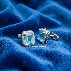 May include: Silver cufflinks featuring rectangular, light blue gemstones, framed by smaller clear stones. The cufflinks rest on a textured, deep blue velvet surface. The text "shaheen enterprises" is visible.