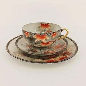 Könnte beinhalten: Ein dreiteiliges Porzellan-Teeservice. Die Tasse, die Untertasse und der Teller haben einen grauen Hintergrund mit roten und orangen Blattmotiven. Die Tasse hat einen goldenen Henkel und einen schwarzen Rand mit Goldverzierungen. Das Set ist wahrscheinlich Vintage.