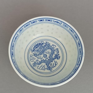 Op de afbeelding: Een witte keramische kom met een blauwe onderglazuur drakenillustratie in het midden. De binnenrand is versierd met een blauw geometrisch randpatroon en de wanden van de kom vertonen een doorschijnend rijstkorrelpatroon over het gehele oppervlak.