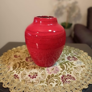 Op de afbeelding: Een glanzende rode vaas met een afgeronde vorm en een licht uitlopende hals. De vaas staat op een decoratief rond kleedje met bloemmotieven in groen, bordeauxrood en goud. De achtergrond is wazig, wat duidt op een interieur.