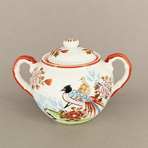 Può includere: Zuccheriera in ceramica bianca con coperchio, con un design colorato di uccelli e fiori. La zuccheriera ha manici e bordi rossi, con un coperchio abbinato. Il design include accenti rosa, rossi, blu e gialli.