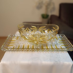 Puede incluir: Un juego de servir de cristal transparente con detalles amarillos. El juego incluye una bandeja rectangular y un cuenco redondo. El cuenco y la bandeja tienen un diseño decorativo. El juego está sobre un mantel blanco.