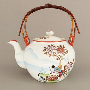 Può includere: Una teiera in ceramica bianca con una scena dipinta a mano di uccelli e fiori colorati, inclusi fiori rosa e rossi. La teiera presenta un beccuccio curvo, un coperchio abbinato e un manico in rattan intrecciato di colore marrone.