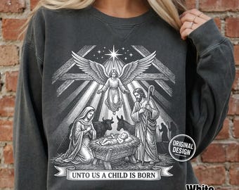 May include: A charcoal gray crewneck sweatshirt featuring a white graphic print of a nativity scene with an angel, Mary, Joseph, a baby in a manger, and animals. Below the scene, a banner contains the text UNTO US A CHILD IS BORN.