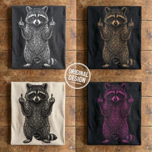 Four folded t-shirts in dark grey, black, cream, and black showing white, beige, black, and pink variations of the edgy raccoon design