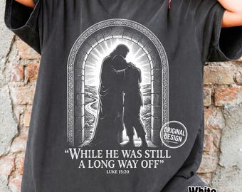 Może przedstawiać: Ciemnoszary t-shirt z białym nadrukiem graficznym przedstawiającym dwie postacie obejmujące się w łukowatym przejściu. Tekst głosi "WHILE HE WAS STILL A LONG WAY OFF" oraz "LUKE 15:20", z małą okrągłą pieczątką "ORIGINAL DESIGN".