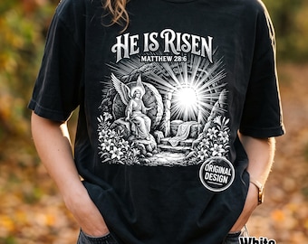 Może przedstawiać: Czarny t-shirt z białą grafiką przedstawiającą anioła siedzącego przy otwartym grobie, z napisem "HE IS RISEN" oraz "MATTHEW 28:6". Wzór zawiera promienie światła wydobywające się z wejścia do grobu oraz kwiaty lilii.