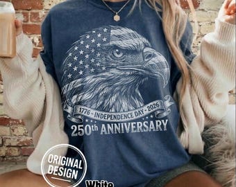 Puede incluir: Una camiseta azul marino con un gráfico de águila blanca que incorpora un diseño de bandera. El texto dice "1776 - Independence Day - 2026" y "250th Anniversary". Incluye "Original Design" y "White".