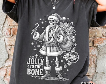 Może przedstawiać: Ciemnoszary t-shirt z białą grafiką przedstawiającą szkielet przebrany za Świętego Mikołaja, trzymający laskę cukrową i worek z prezentami. Napis głosi JOLLY TO THE BONE. Wzór zawiera gwiazdki, płatki śniegu oraz okrągłą pieczęć z napisem ORIGINAL DESIGN.