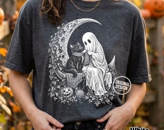 Może przedstawiać: Ciemnoszary melanżowy t-shirt z białą grafiką przedstawiającą ducha siedzącego na półksiężycu i robiącego na drutach szalik dla czarnego kota. Wzór zawiera liście bluszczu, dynię oraz kłębek wełny z okrągłą odznaką w prawym rogu.